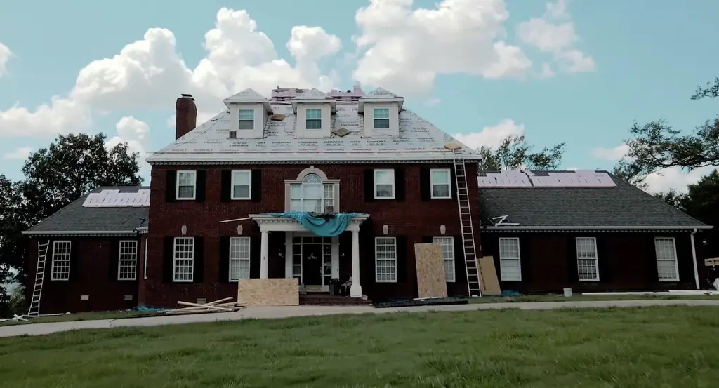 Large brick colonial mid-job with the main roof stripped to white underlayment and ladders against the dormers