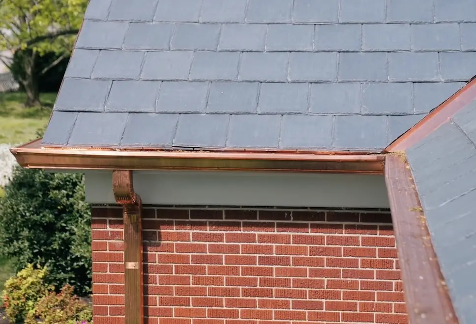 Grey slate roof edge with a copper gutter and downspout on red brick